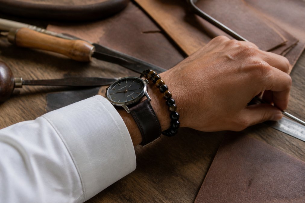 Man's wrist wearing beaded onyx and tiger eye bracelets with a leather watch and white shirt cuff