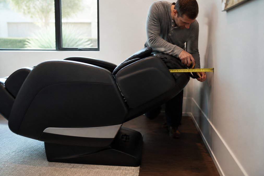 Person using tape measure to check distance between reclined massage chair backrest and wall, showing the gap during full recline in a modern living room with natural lighting