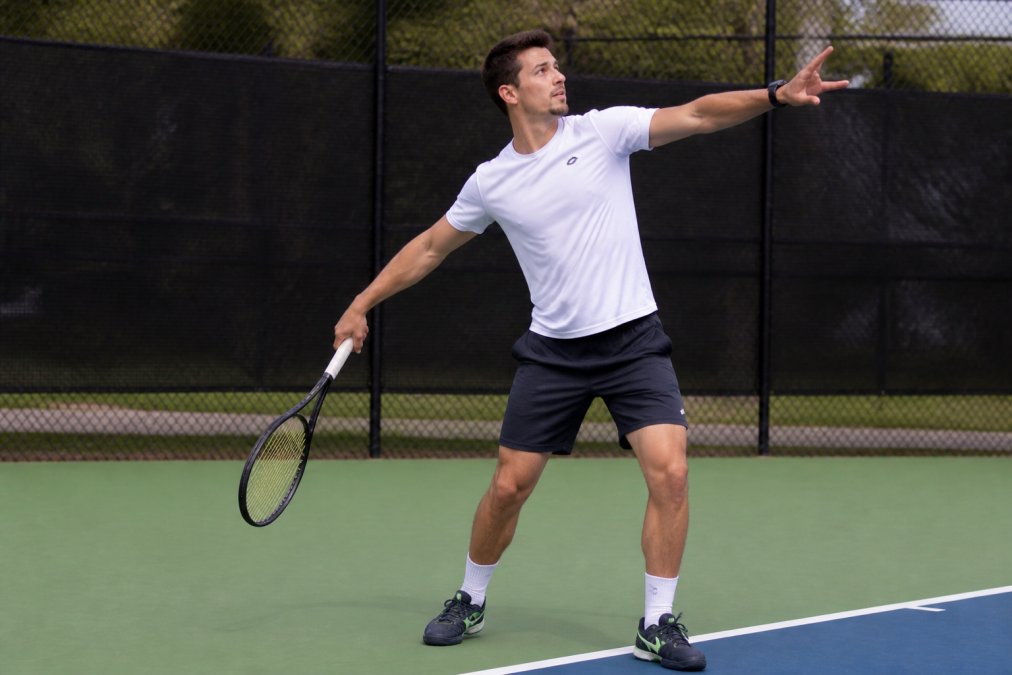 Player in trophy position at the start of serve motion, showing clear shoulder rotation, extended non-dominant arm for balance, and racket head below hip level, captured in bright outdoor court lighting