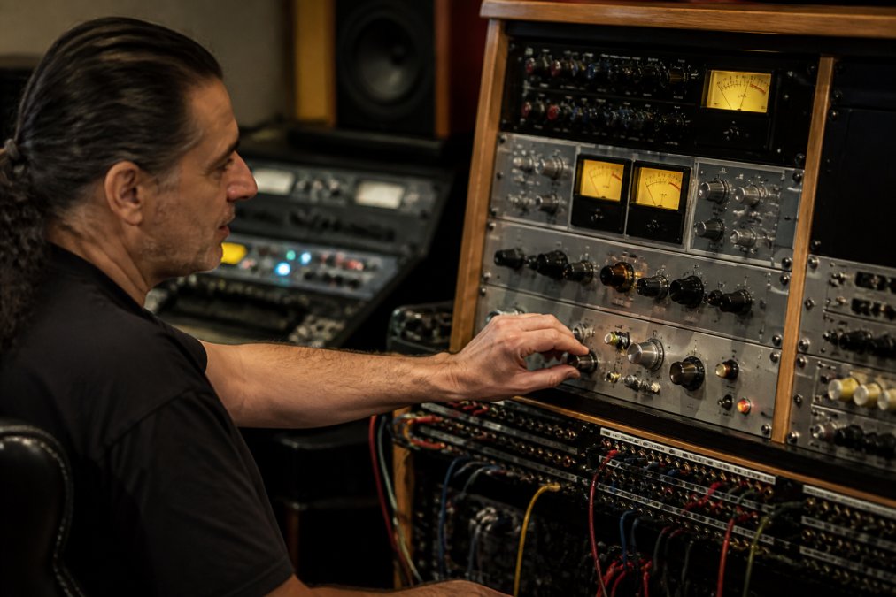 A professional mastering studio rack filled with analog outboard gear, including a compressor and equalizer, with glowing VU meters and patch bay cables visible