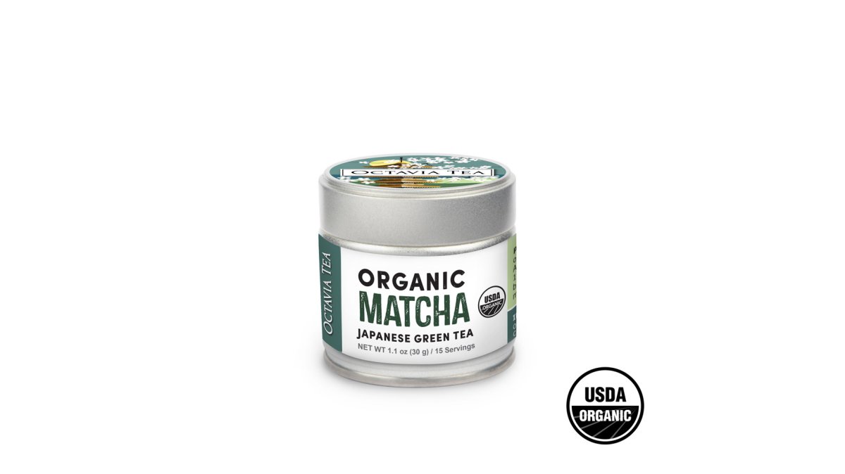 A ceramic matcha bowl filled with vibrant green matcha powder beside a bamboo whisk, with a glass teapot of steeped green tea leaves behind it on a wooden table in soft natural light
