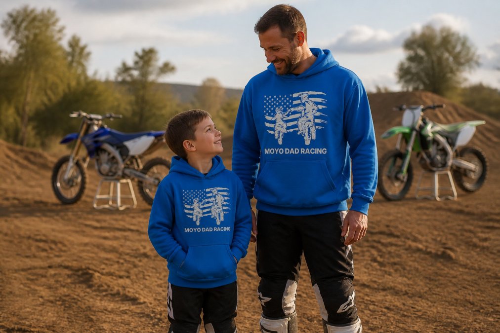 Father and young son wearing matching motocross-themed hoodies and racing jerseys standing together at a dirt track with motorcycles visible in the background, natural afternoon lighting