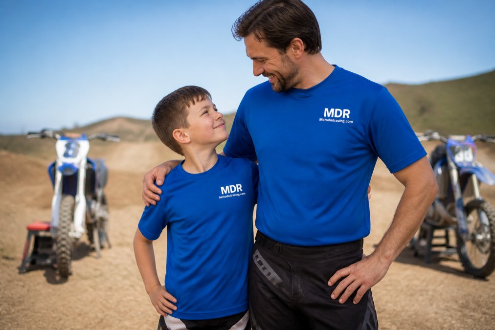 A father and young son wearing matching motocross-themed jerseys standing together at a dirt bike track, both smiling with bikes visible in the background