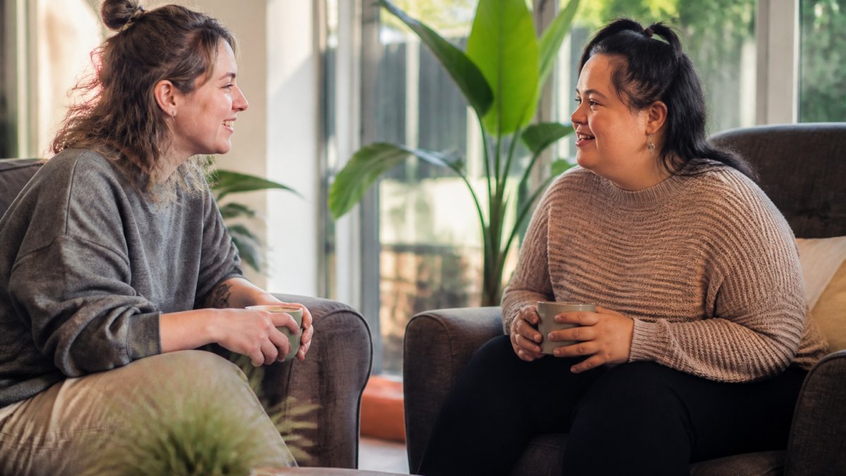 Two people sitting together in a comfortable living room, having a relaxed conversation over tea or coffee, natural afternoon light streaming through windows