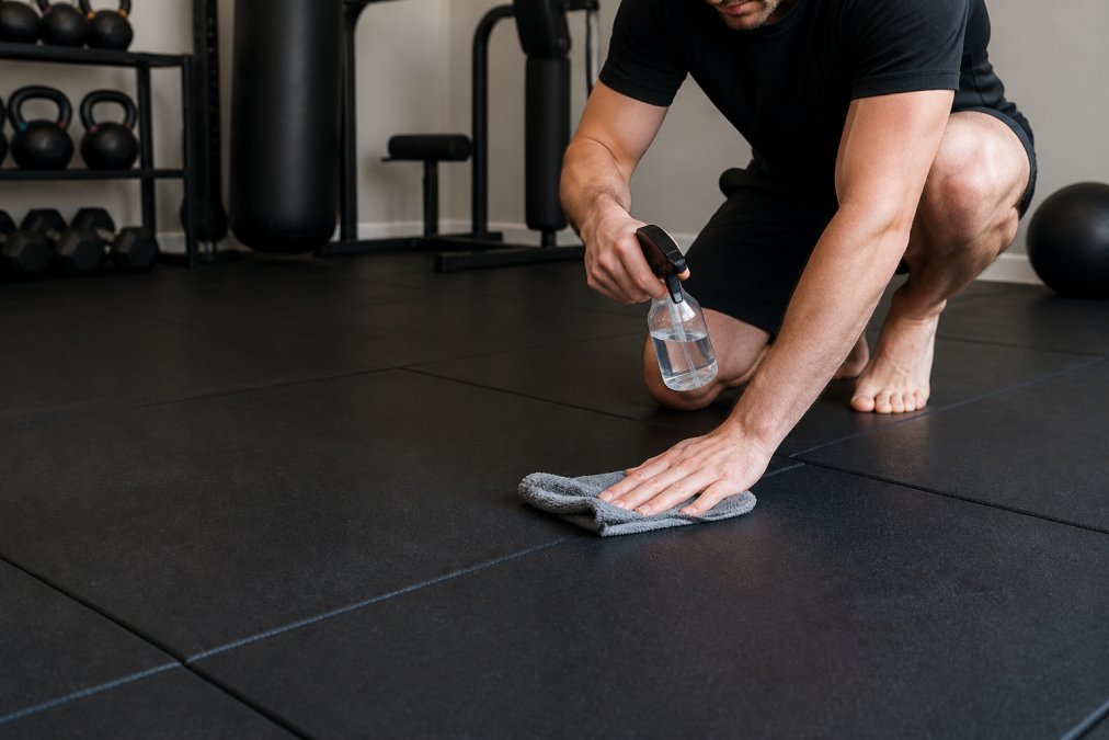 A home gym mat being cleaned with a spray bottle of sanitizing solution and microfiber cloth, showing proper technique for disinfecting the surface, with equipment visible in background