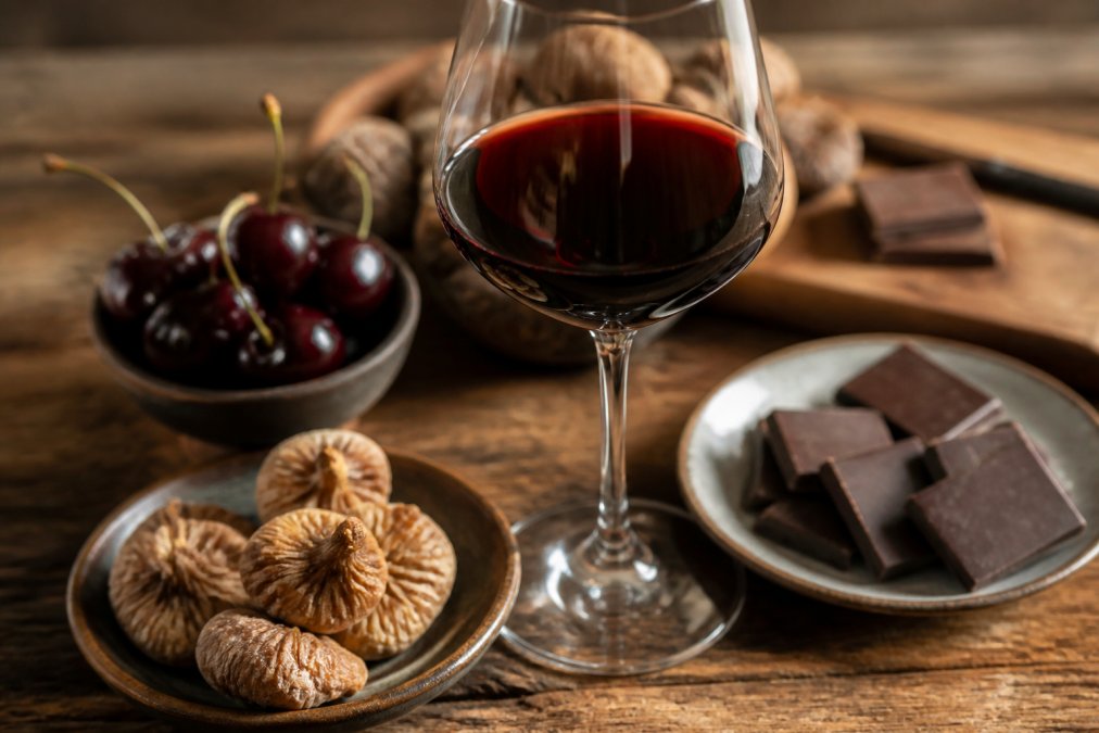 Nahaufnahme eines tiefrubinroten Glases Mavrodaphne-Wein auf einem Holztisch, umgeben von getrockneten Feigen, dunklen Kirschen und einem Stück dunkler Schokolade, warmes natürliches Licht