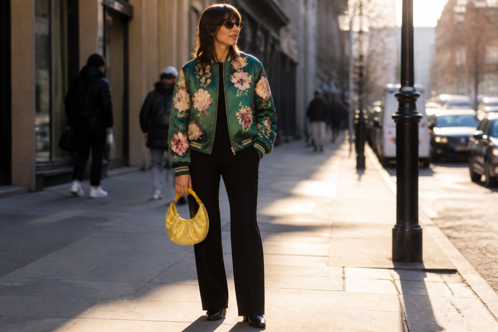A fashion-forward woman in a bold satin bomber jacket covered in floral appliqué, paired with straight-leg black trousers and simple black shoes, holding a rhinestone bag, photographed on a sunlit city street with blurred urban background