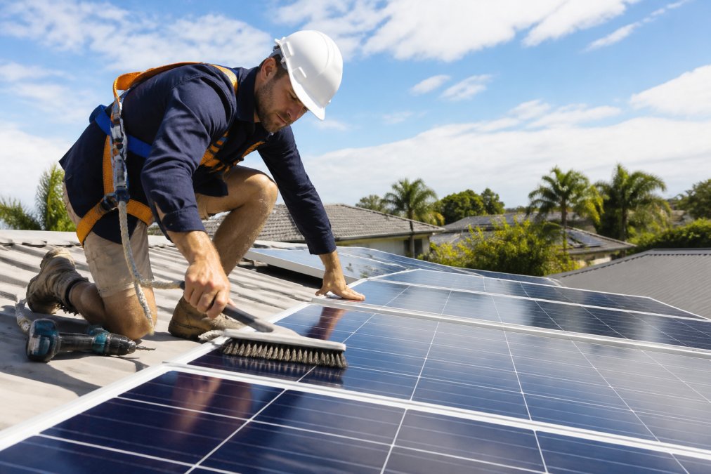 Professional technician in safety harness performing maintenance on rooftop solar panels, cleaning panels with soft-bristle brush and checking electrical connections in bright daylight, residential home setting