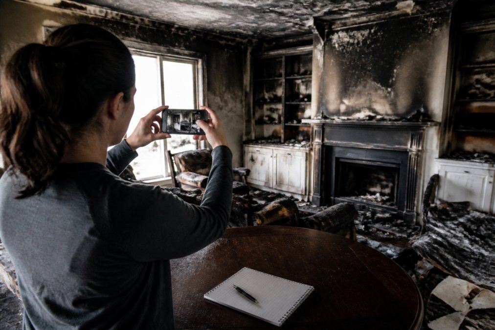 A homeowner photographing fire-damaged belongings and structural damage in a residential room, using a smartphone, with a notepad and pen nearby