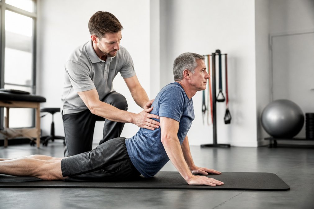 A physical therapist in a bright, modern clinic guiding a middle-aged male patient through a prone press-up exercise on a mat, demonstrating proper hand placement and form