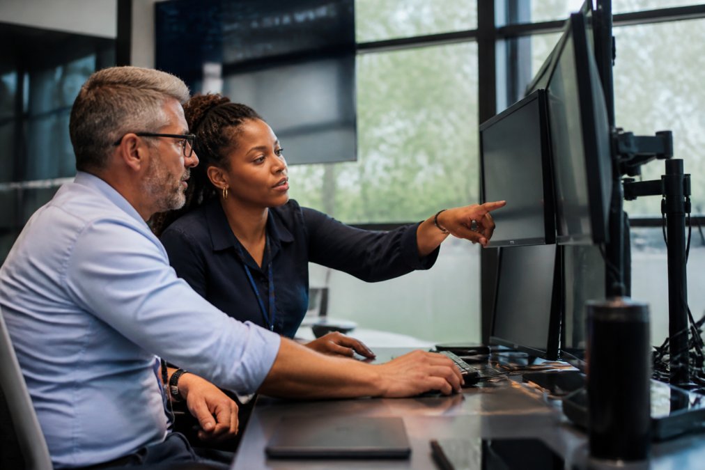 Healthcare IT director and CISO reviewing security monitoring dashboard on multiple screens in a modern SOC environment with natural lighting