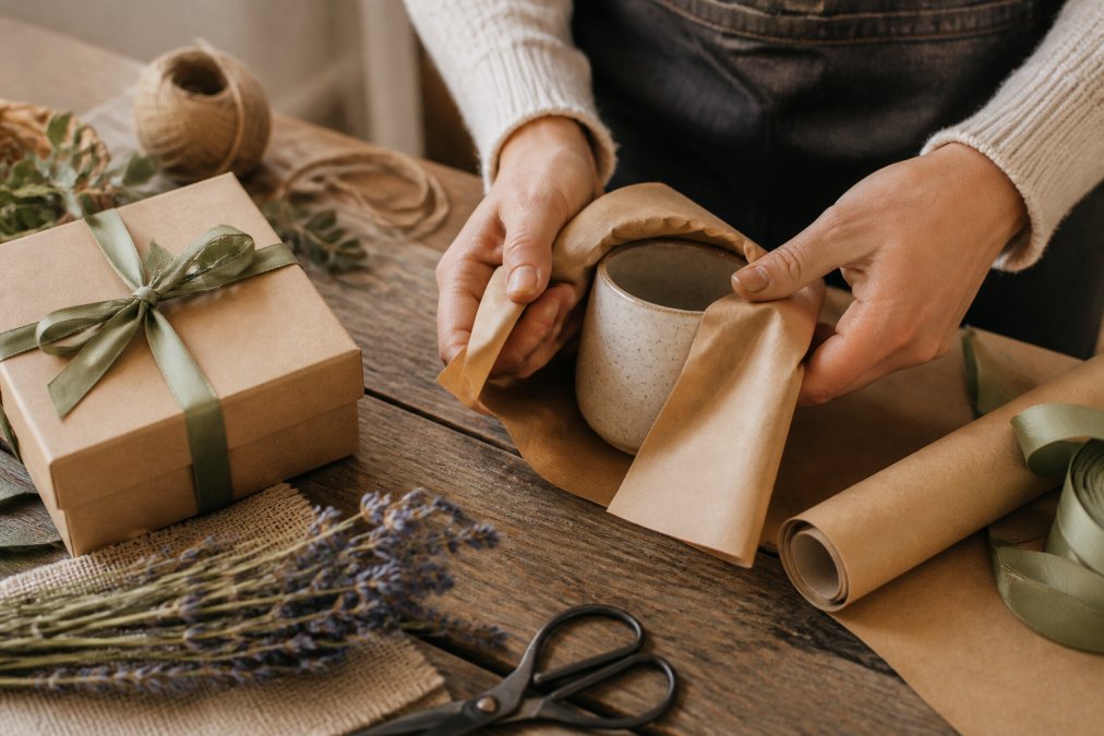 Hands carefully wrapping a ceramic mug in sustainable kraft paper with natural twine, artisan gift box with eco-friendly materials and ribbon on wooden table, thoughtful presentation