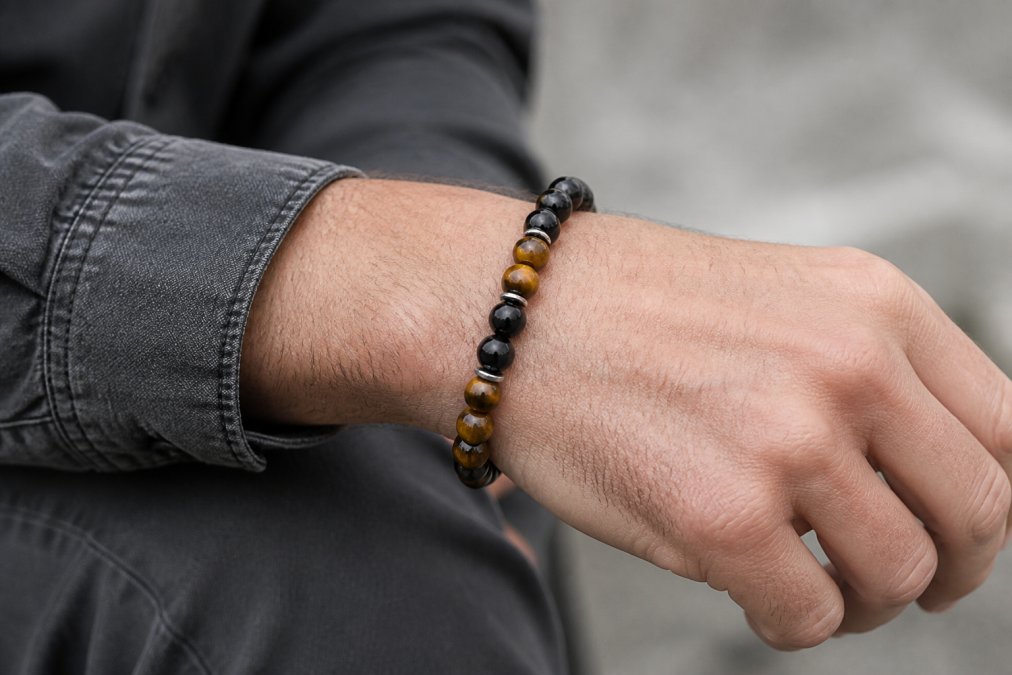 Close-up of a man's wrist wearing a natural stone bracelet in black onyx and tiger eye, photographed in natural daylight with neutral background showing stone details and masculine aesthetic