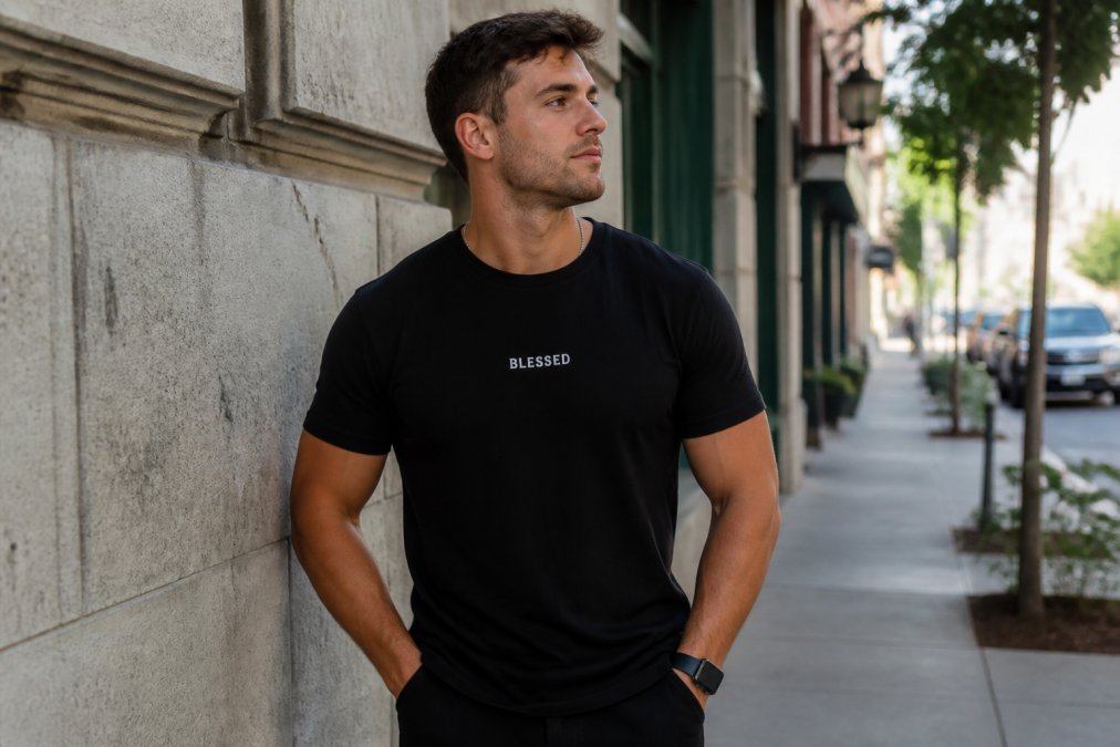 A young man in his late 20s wearing a simple black t-shirt with small white text reading 'BLESSED', standing in an urban street setting with soft natural light, looking confident and approachable