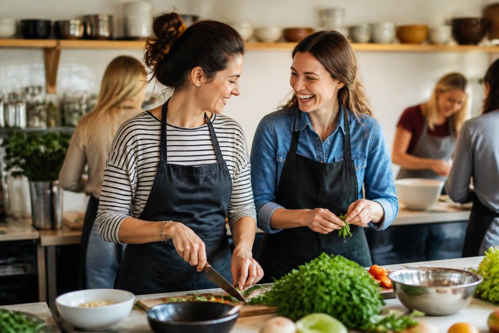 Two adult women enjoying a cooking class together in a bright, modern kitchen studio, laughing while preparing food, creating a shared memorable moment