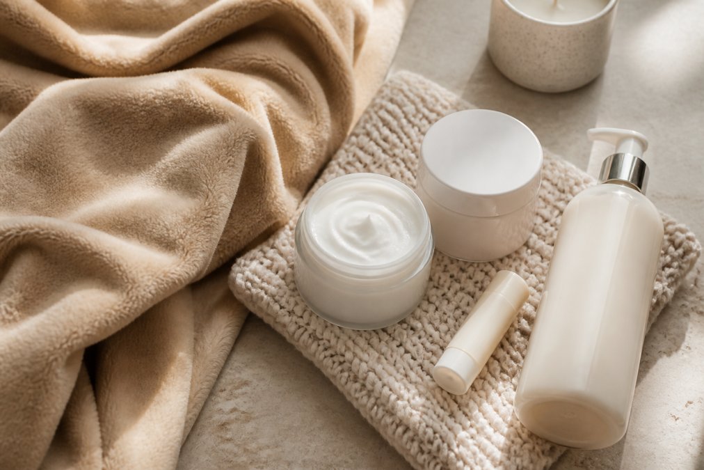 Flat lay of cozy self-care items including soft fleece blanket, unscented moisturizer jar, lip balm, and luxurious body lotion arranged on a warm neutral surface with soft natural lighting