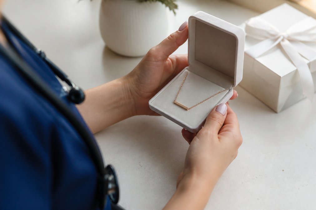 Close-up of a nurse's hands holding a small personalized name necklace in a soft-touch gift box on a clean table, warm natural light