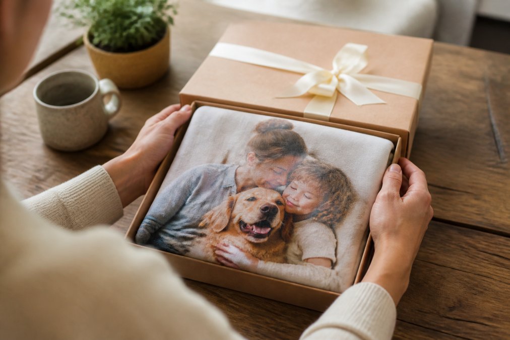 Close-up of an adult's hands holding a beautifully wrapped personalized keepsake gift box with a custom photo blanket partially unfolded, soft warm lighting on a wooden table