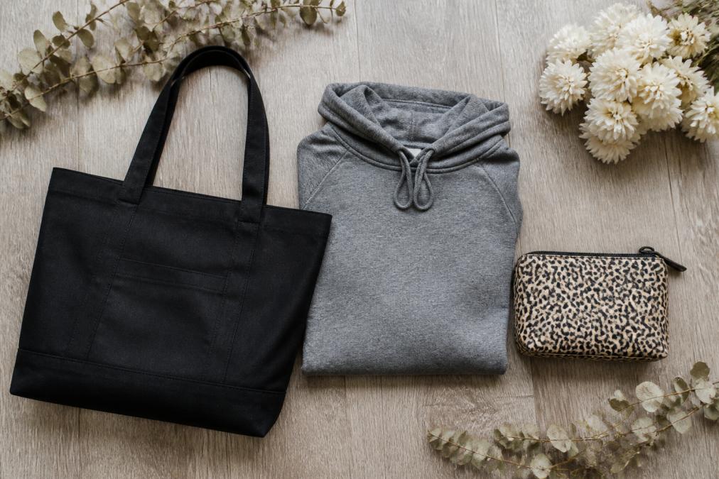 Flat lay of stylish women's accessories arranged on a light wood surface, including a deep black canvas tote bag with gold lettering, a neatly folded heather-grey hoodie, and a small animal print cosmetic pouch, surrounded by dried eucalyptus and white flowers, photographed from directly above in soft natural light