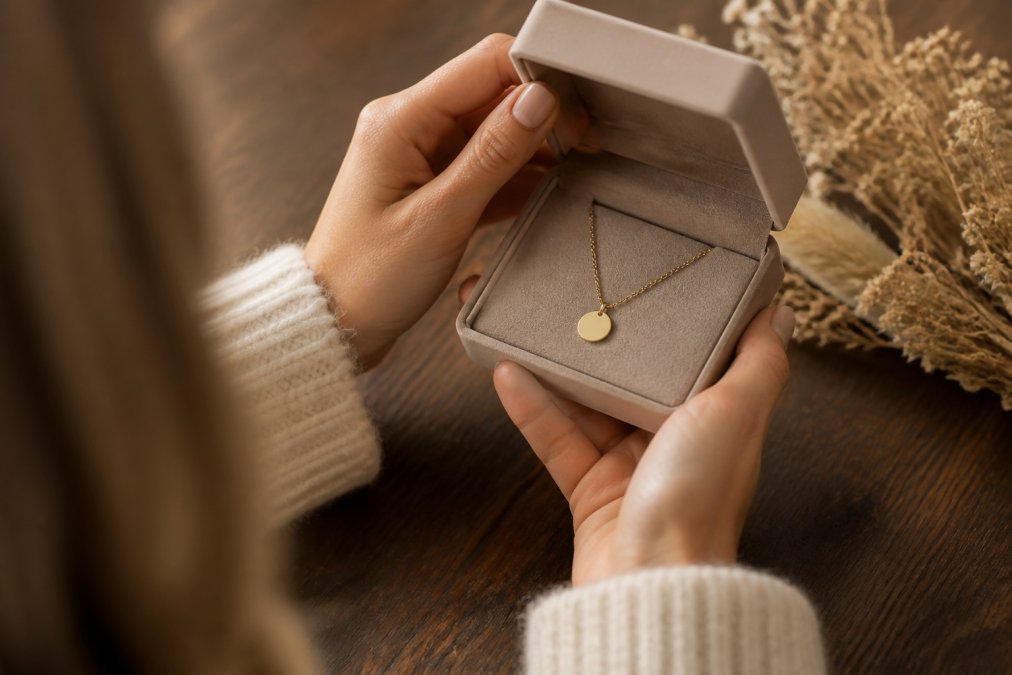 Close-up of a woman's hands opening a soft-touch jewelry box to reveal a delicate gold pendant necklace, warm natural light on a wooden table with dried flowers nearby