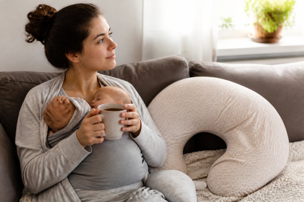 A tired parent sitting on a sofa in soft morning light, holding a warm mug with both hands while a baby sleeps in a sling carrier against their chest, a nursing pillow propped beside them