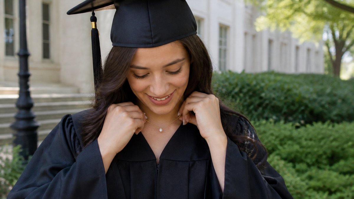Young woman in graduation cap and gown smiling while adjusting a delicate gold necklace, natural outdoor lighting