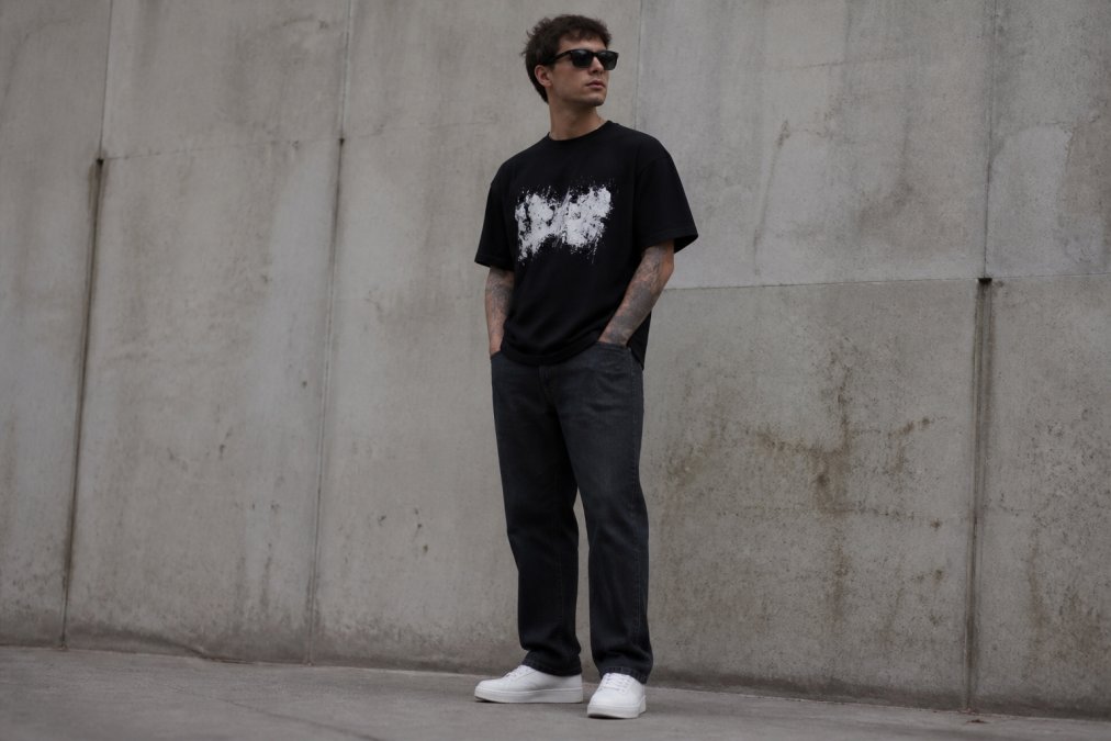A person wearing a heavyweight black graphic tee with a bold white chest print, styled with dark straight-leg denim and plain white sneakers, standing against a concrete wall in soft overcast daylight