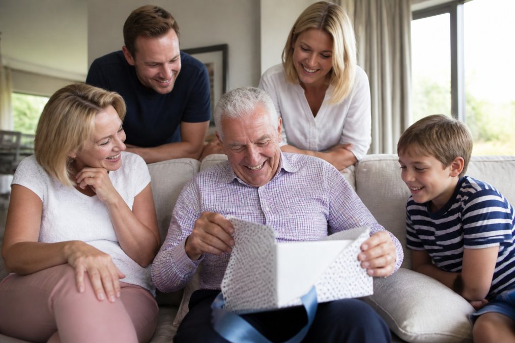 An older adult opening a beautifully wrapped gift with a warm, genuine smile while surrounded by family members watching with affection in a bright, comfortable living room