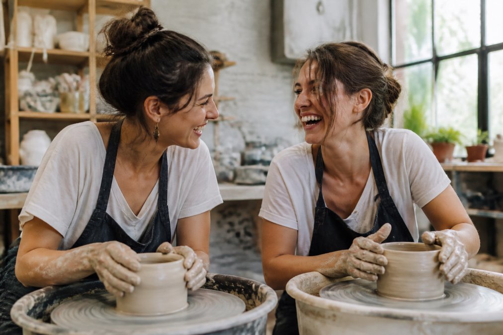Two friends laughing while working with clay in a pottery studio, a perfect example of shared non-material gifts.