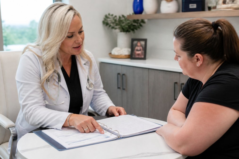 Board-certified physician consulting with patient in clinical setting, reviewing personalized treatment documents and metabolic health markers on tablet, modern medical office with natural lighting