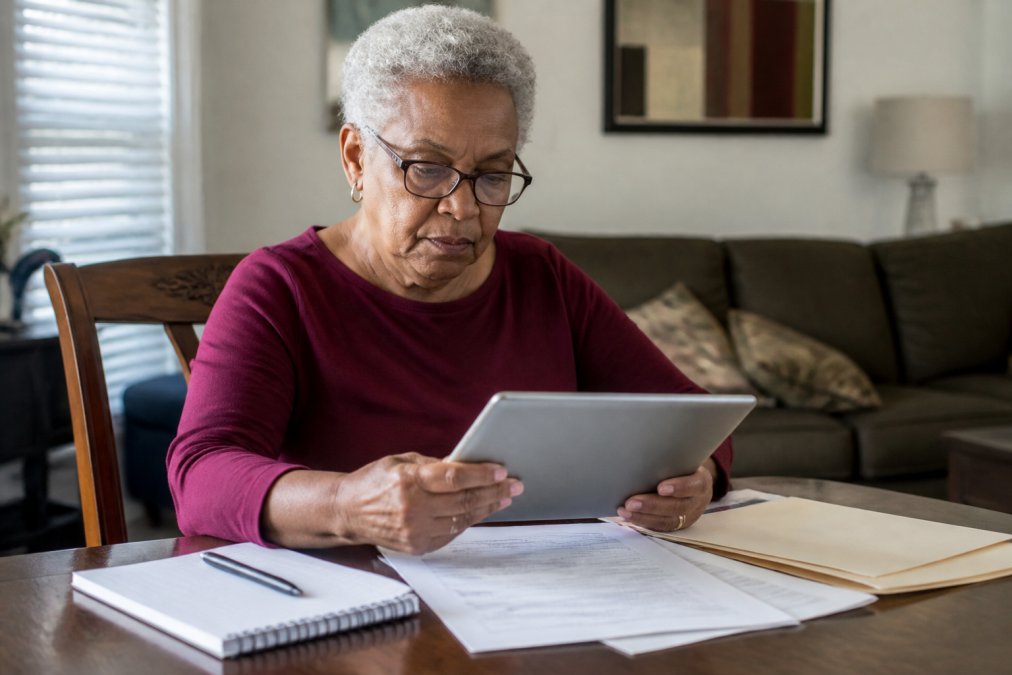 Senior adult at home reviewing Medicare documents and enrollment forms on a tablet, with a notebook and pen on the table