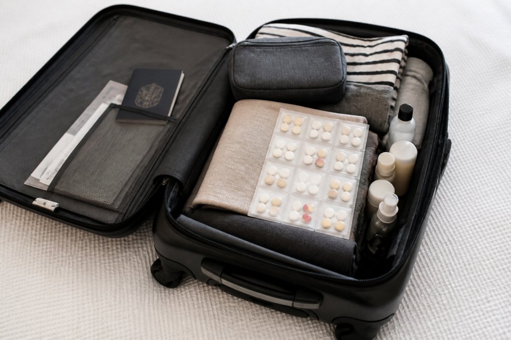 Traveler's open suitcase showing neatly organized pre-sorted medication packets alongside passport, travel documents, and toiletries on a light background