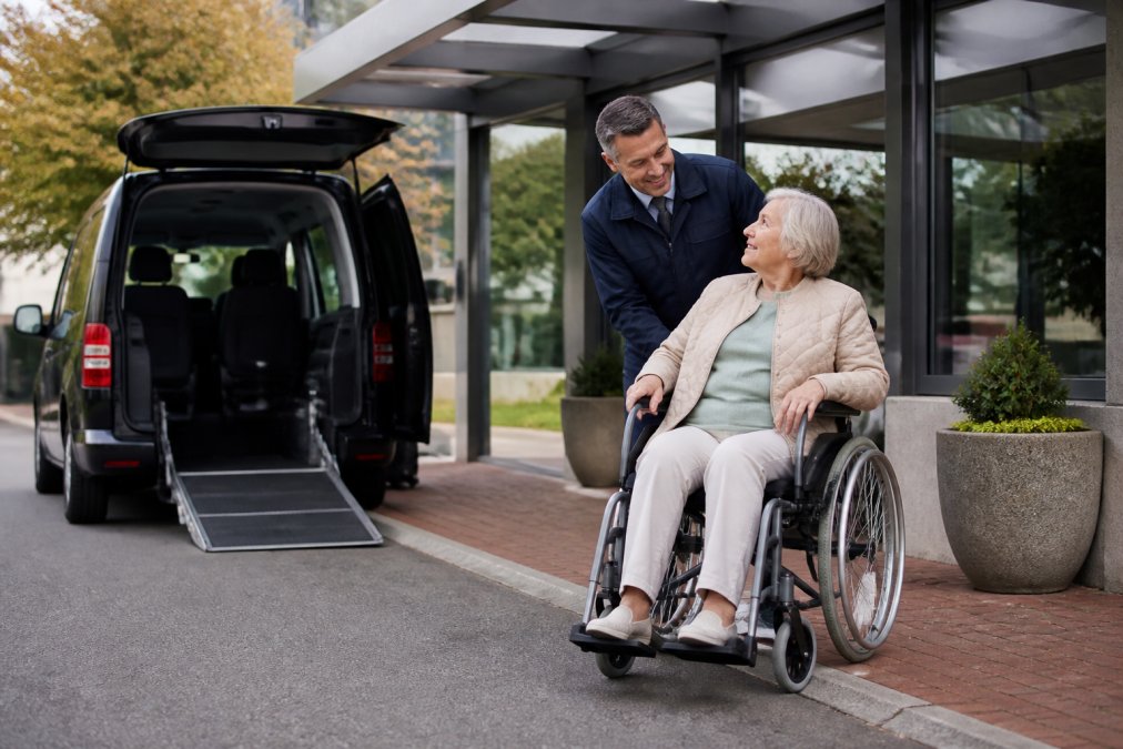 Oudere rolstoelgebruiker die wordt geholpen door een vriendelijke chauffeur in uniform bij de ingang van een medische kliniek, met een toegankelijke taxi zichtbaar op de achtergrond in zachte daglichtomstandigheden