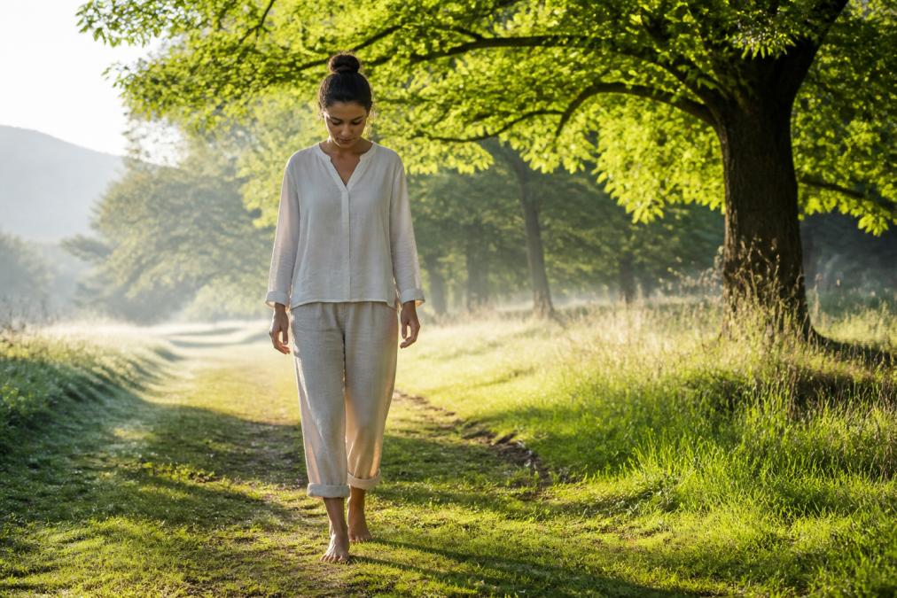 A person walking barefoot on a grassy path outdoors on a sunny morning, looking down mindfully at the ground, arms relaxed at their sides, soft natural light filtering through trees overhead