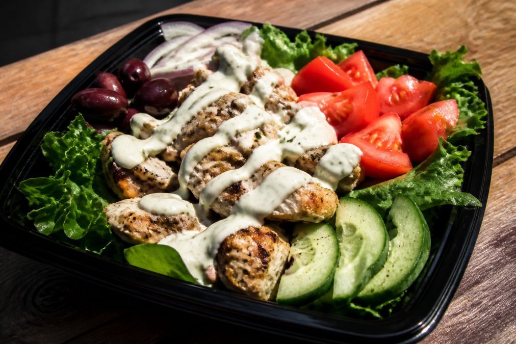 Close-up of a freshly prepared Mediterranean takeout bowl with grilled chicken, vibrant leafy greens, ripe tomatoes, cucumber slices, kalamata olives, and creamy tzatziki sauce drizzled on top, with natural daylight illuminating the fresh ingredients
