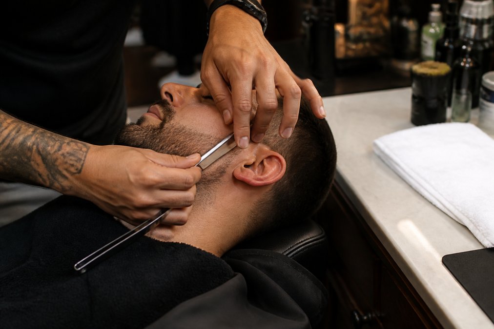 Gros plan des mains d'un barbier tenant un rasoir droit et traçant le contour net de barbe d'un client, avec une serviette chaude blanche posée sur le comptoir en marbre à côté, éclairage de salon chaud