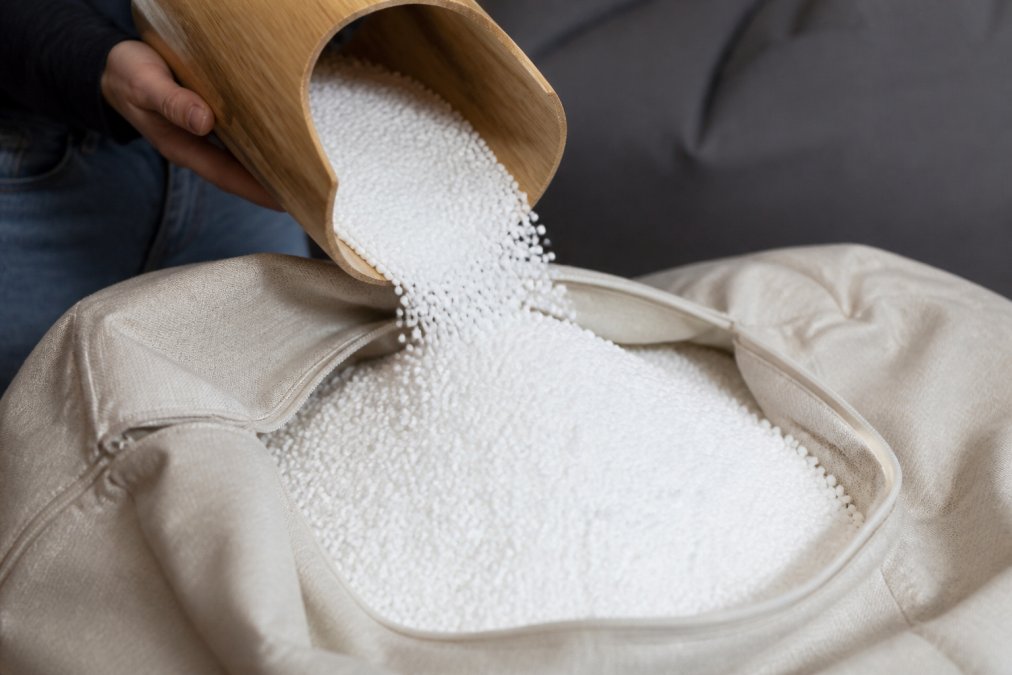 Gros plan sur des billes de polystyrène EPS blanc brillant versées doucement dans un pouf beige, montrant la texture légère et granulaire du matériau, avec une main tenant un entonnoir en bois
