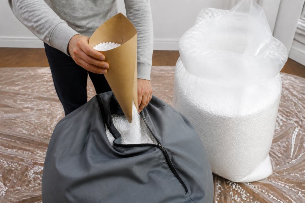 Personne versant du remplissage dans un pouf à l'aide d'un entonnoir en carton, une main tenant fermement l'entonnoir tandis que l'autre guide le flux de billes, dans un espace bien éclairé avec un sol protégé par du plastique transparent