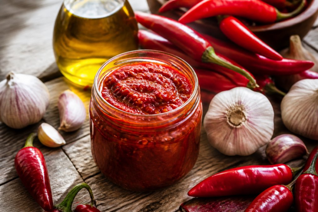 Close-up of fresh red peppers and garlic cloves surrounding an open jar of vibrant red artisanal harissa paste on rustic wooden surface with olive oil in background, natural lighting