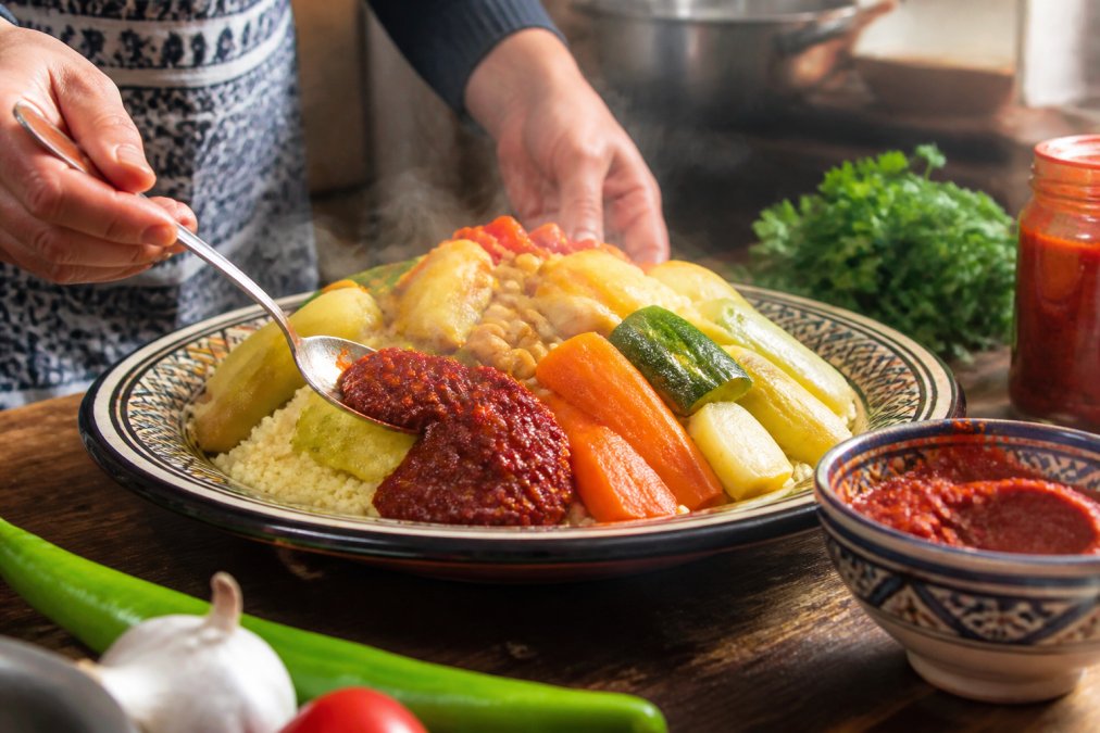 Plate of steaming couscous with colorful vegetables and a spoonful of red harissa paste applied to the side, hands preparing the dish, warm culinary ambiance with natural kitchen lighting