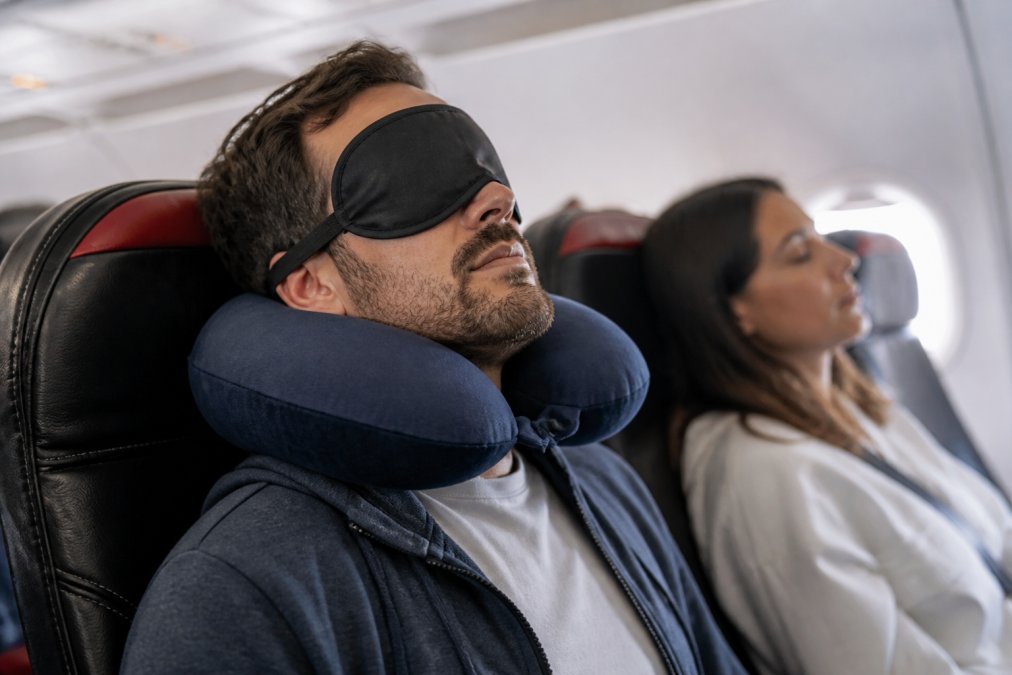 Voyageur endormi confortablement en avion, la tête reposant sur un coussin cervical ergonomique, masque de sommeil occultant en place, montrant le maintien correct de la nuque et du cou dans une position neutre