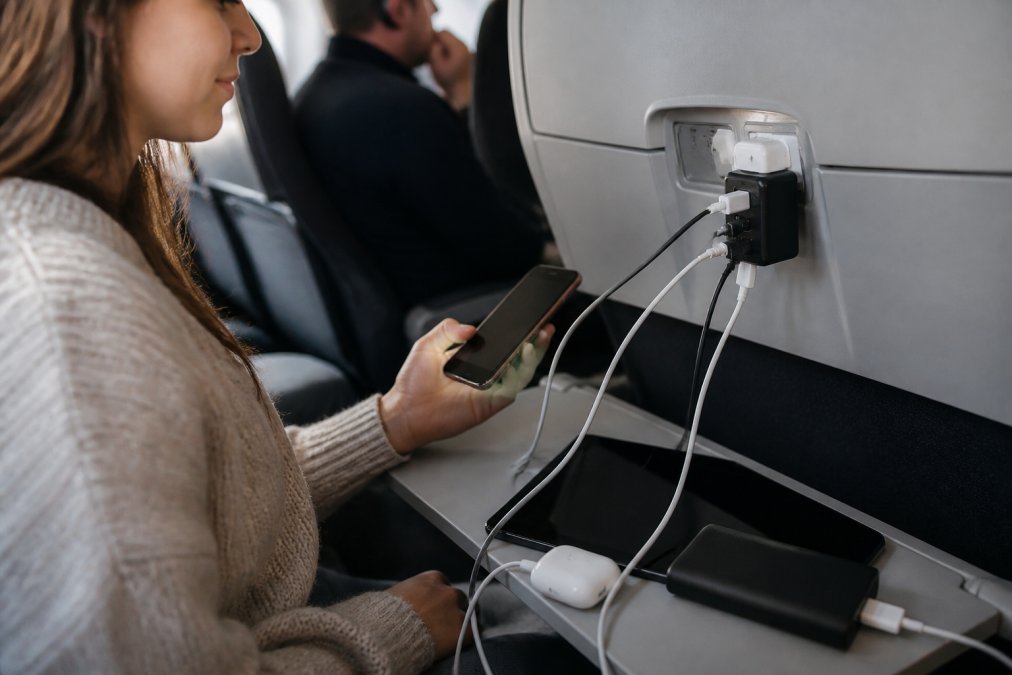 Adaptateur universel branché dans une prise d'avion avec plusieurs appareils (téléphone, tablette, écouteurs sans fil) connectés simultanément, montrant la charge multiple en action dans une cabine d'avion