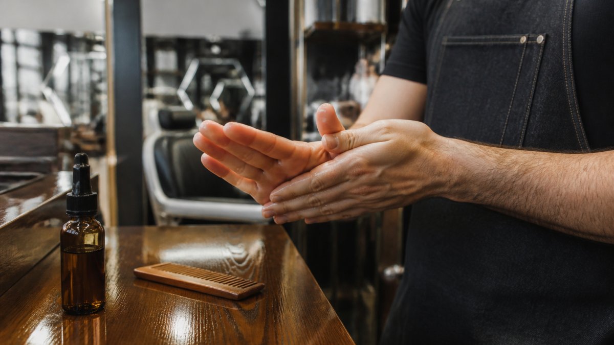 Close-up of a man's hands warming a few drops of beard oil between his palms in a bright barbershop, with a wooden comb and amber glass bottle on the counter nearby