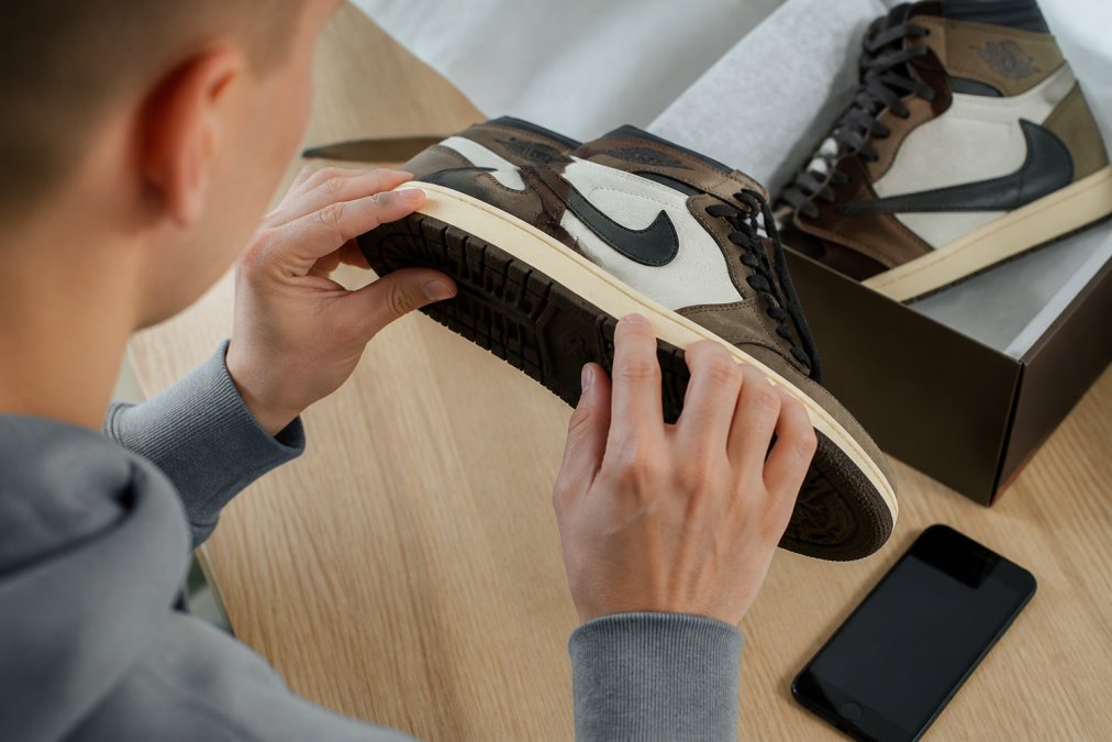 Person's hands carefully inspecting the sole and stitching of a sneaker under bright light, with a smartphone nearby showing an authentication app