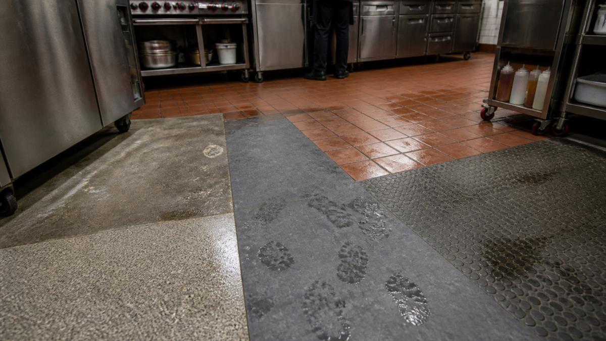 Close-up view of a busy commercial kitchen floor with multiple flooring material samples visible, showing wear patterns, footprints, and water marks under bright overhead lighting in a restaurant setting