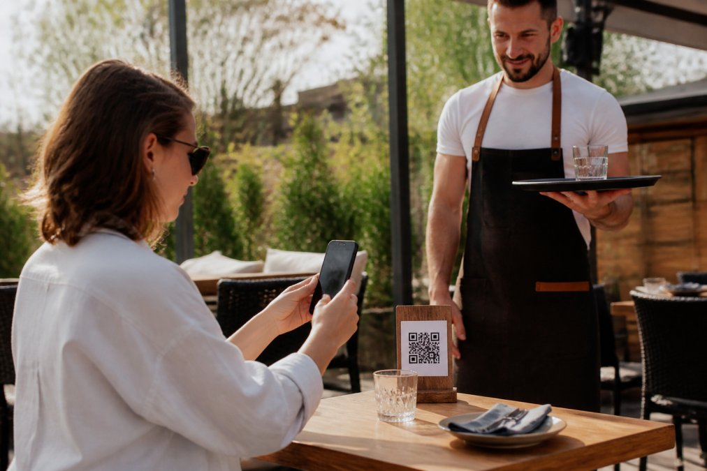 Cliente en un restaurante escaneando un código QR con su smartphone mientras un camarero espera junto a la mesa con una bandeja, luz natural cálida de mediodía