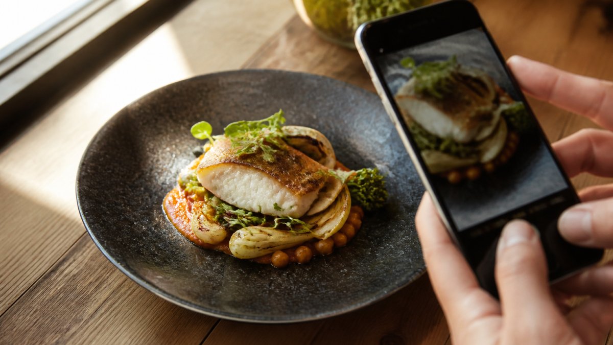 Close-up de un plato gourmet fotografiado desde ángulo de 45 grados con luz natural suave entrando desde la izquierda, mostrando texturas y colores vibrantes del alimento, con un smartphone en el borde del encuadre capturando la imagen