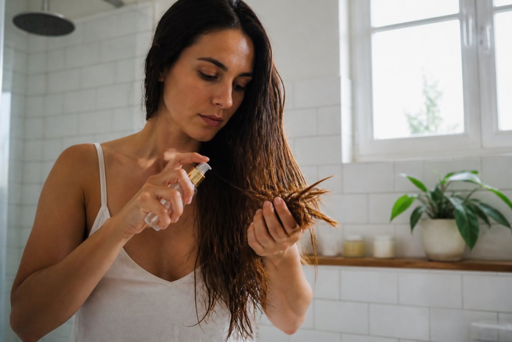 Mujer aplicando uno de los mejores tratamientos veganos para puntas abiertas en su cabello largo dentro de un baño iluminado