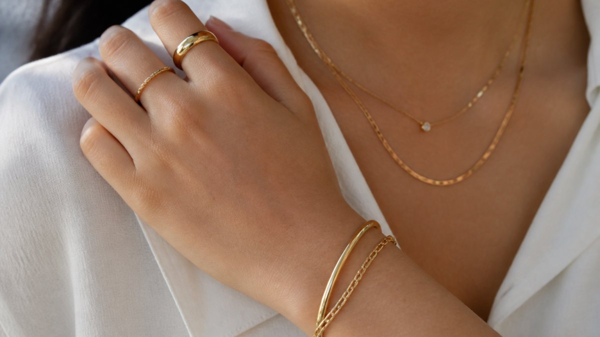 Close-up of a person's wrist and hand wearing layered gold-plated jewelry including a delicate necklace, stacked rings, and a cuff bracelet in natural daylight, showing how pieces coordinate together