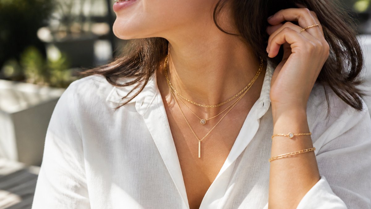 Woman wearing layered delicate gold necklaces and bracelets in natural daylight, showcasing how fine jewelry pieces stack together for everyday wear